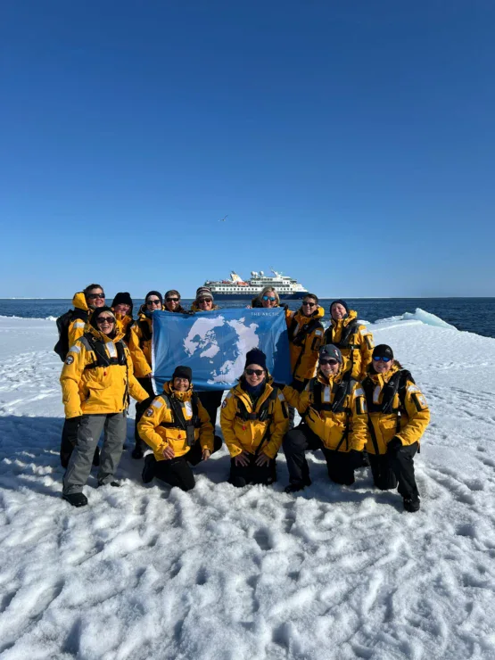 A group of people in yellow jackets pose with a blue map flag on snow, with a ship in the background under a clear blue sky.