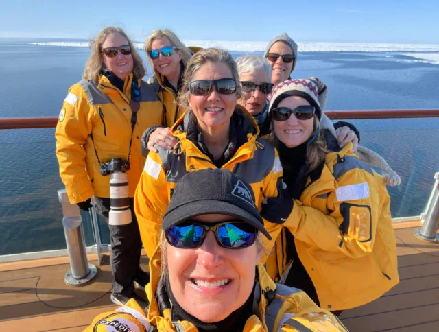 Seven people in yellow jackets pose for a group selfie on a ship deck with calm water and ice in the background on a sunny day.
