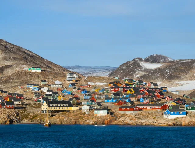 A coastal village with colorful houses sits on rocky terrain, surrounded by snow-dusted hills and bordered by blue water under a clear sky.