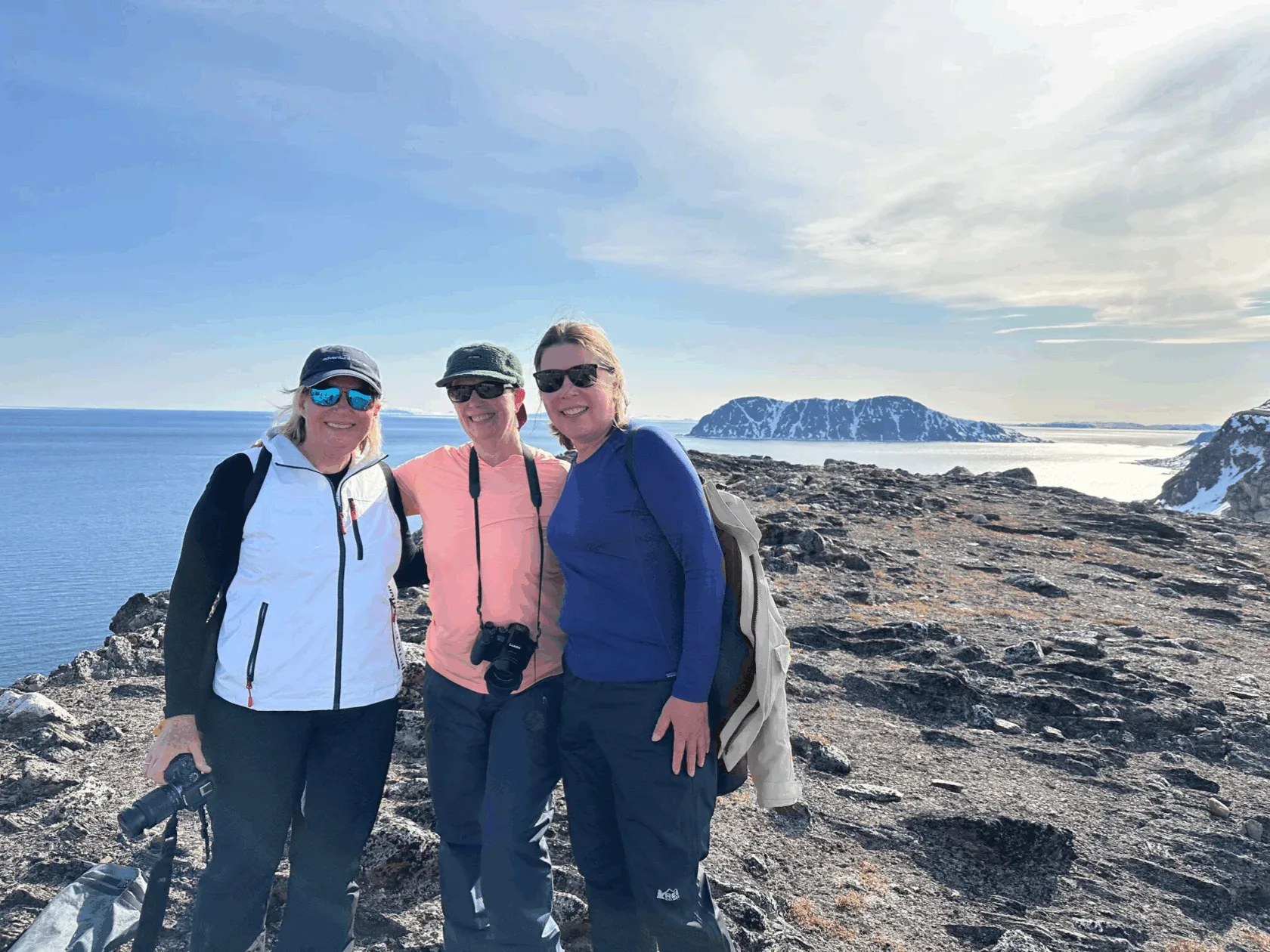 Three women in outdoor gear stand on a rocky coastline with binoculars, smiling at the camera. A distant mountain and body of water are visible under a clear sky.