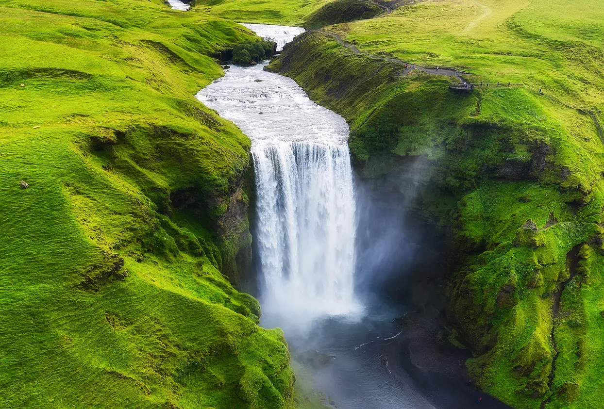 A tall waterfall cascades over a cliff surrounded by lush green hills, with water mist rising from the base.