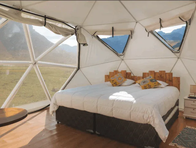 A dome-shaped glamping tent with a double bed, wooden headboard, pillows, nightstand, and large windows overlooking a scenic mountain landscape.