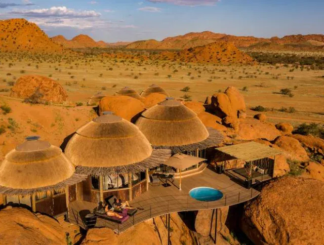 A lodge with thatched-roof huts and a small pool is built among red rocks in a desert landscape with scattered shrubs and distant hills.