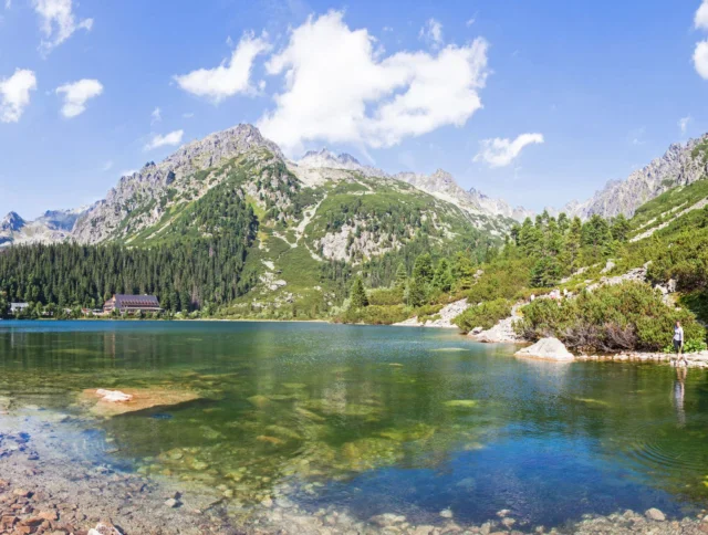 Clear mountain lake with rocky shoreline, surrounded by pine trees and mountains under a blue sky with scattered clouds. A building is visible in the distance near the water’s edge.