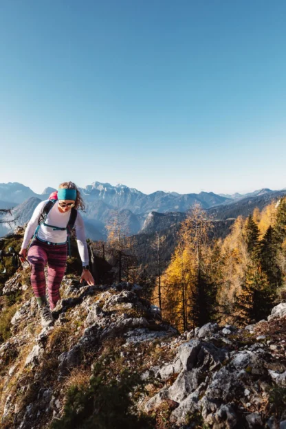 A person wearing hiking gear climbs a rocky mountain trail surrounded by autumn trees, with distant mountains visible under a clear blue sky.