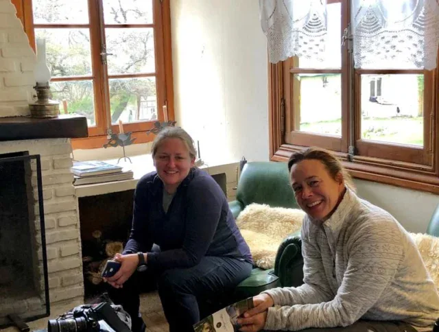 Two people are sitting on a green couch in a cozy room near a fireplace, smiling at the camera. A camera and books are on the table in front of them.