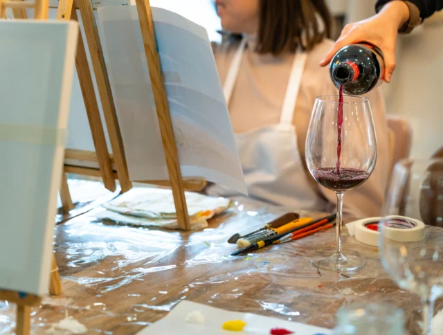 A person pours red wine into a glass on a table with paintbrushes, paint, and easels, suggesting a painting and wine activity.