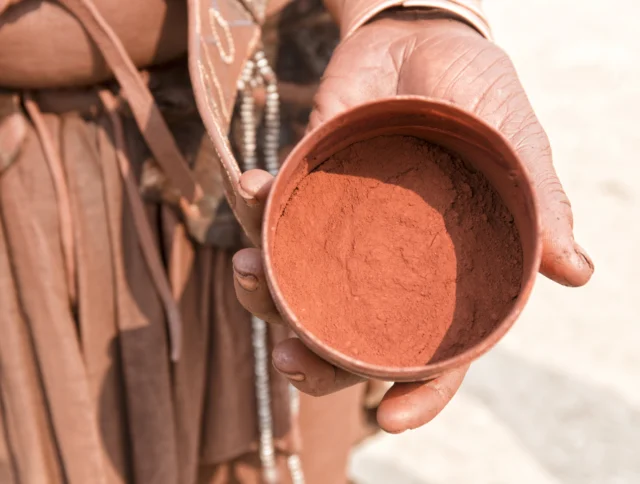 A person holds a small bowl filled with reddish-brown powder, wearing earth-toned clothing and accessories.