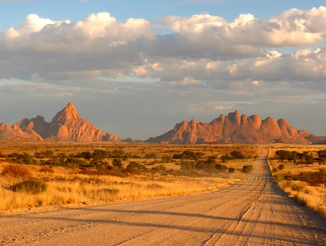 A dirt road stretches through dry grassland toward rocky mountains under a partly cloudy sky at sunset.