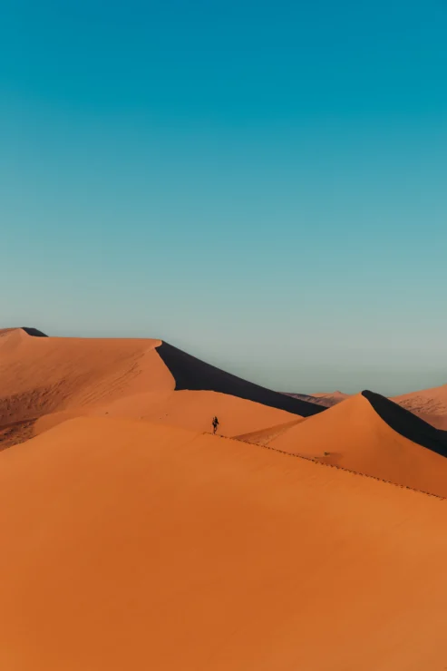 A person walks along the crest of tall orange sand dunes under a clear blue sky in a desert landscape.