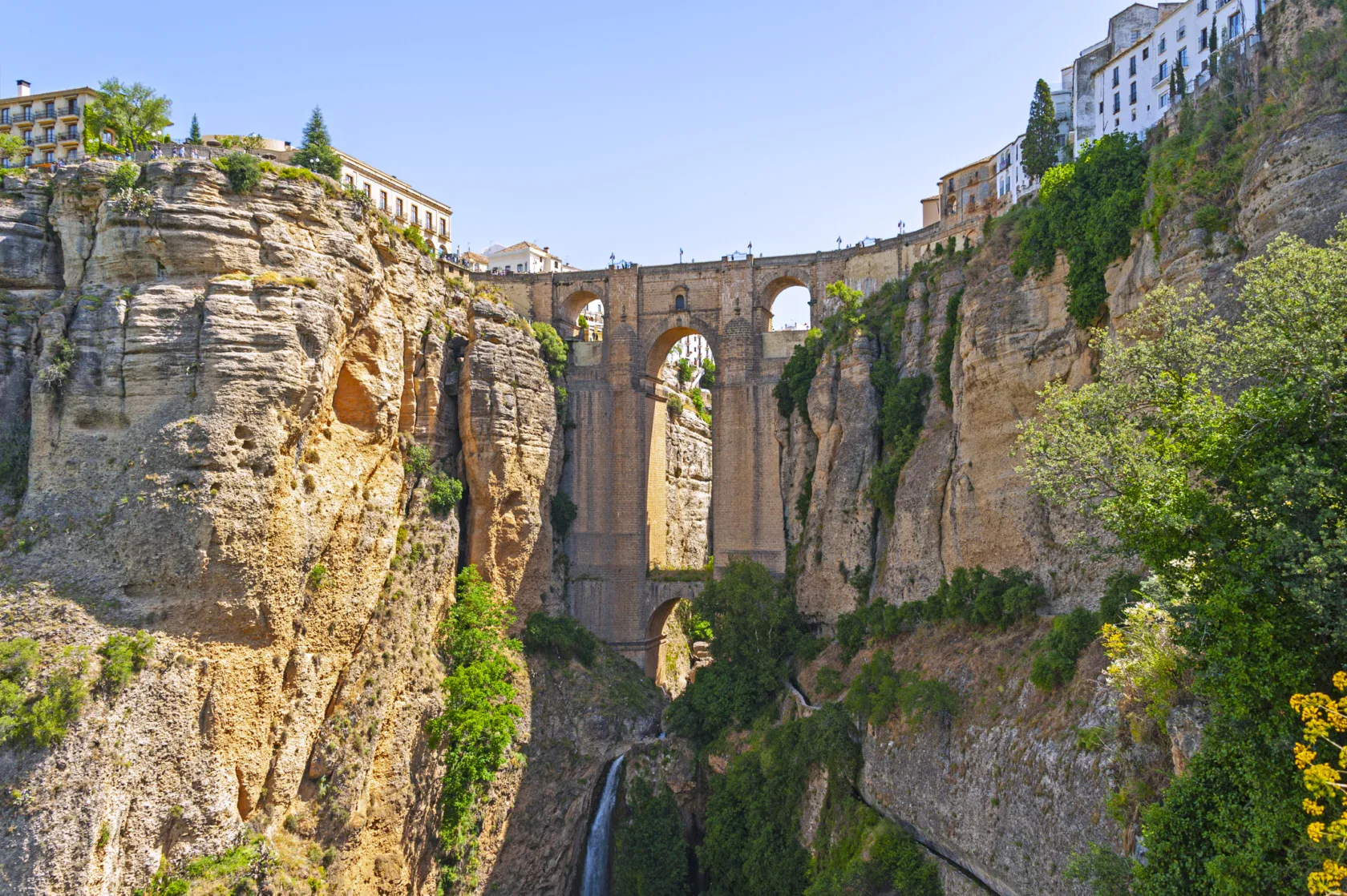 A large stone bridge spans a deep gorge with tall cliffs, connecting buildings on either side; greenery grows on the rocky walls below.