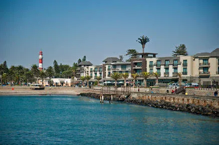 A waterfront scene with modern buildings, palm trees, a rocky shoreline, and a red-and-white striped lighthouse in the background under a clear blue sky.