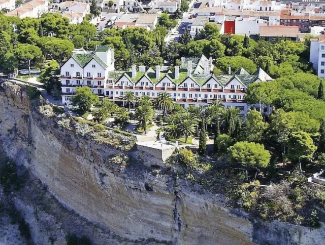 A row of white buildings with green roofs sits atop a steep cliff, surrounded by trees, with a town visible in the background.