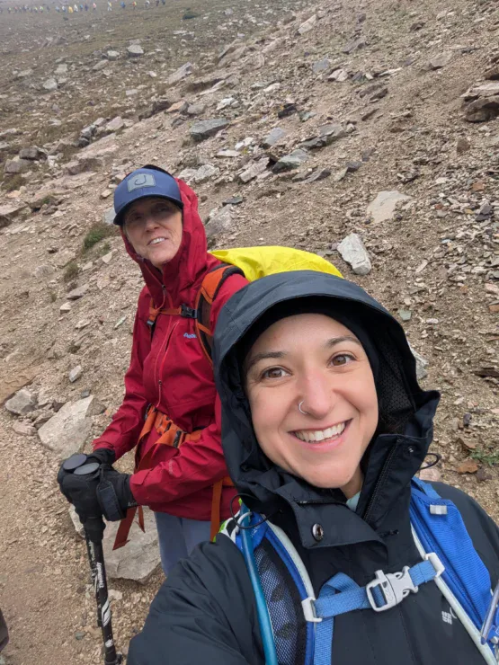 Two people in outdoor gear and backpacks stand on a rocky trail, smiling at the camera. The surroundings are barren with small rocks and patches of grass.