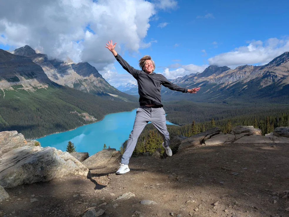 A person jumps in the air on a rocky overlook with a turquoise lake and mountains in the background under a partly cloudy sky.