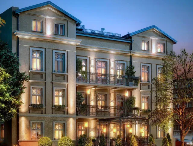A three-story, elegant building with balconies and decorative lighting at dusk, surrounded by trees and bushes, with cars and street signs visible nearby.