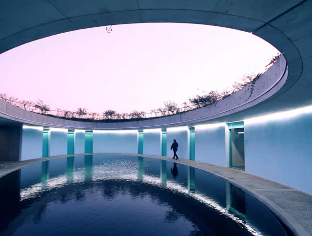 A person walks along a curved concrete path surrounding a circular reflecting pool under a large open skylight with a pale sky.