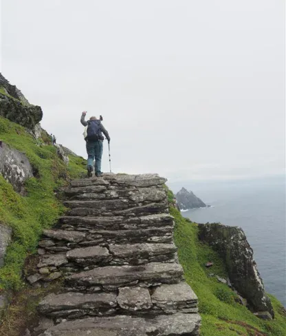 A person with a backpack climbs stone steps on a cliffside path above the ocean under a cloudy sky.