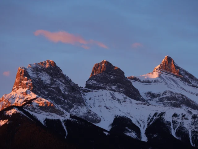 Snow-covered mountain peaks, known as the Three Sisters, are illuminated by the warm light of sunset with a few clouds in the sky.