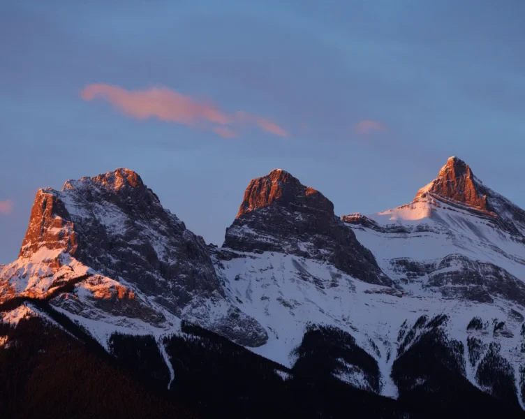 Snow-covered mountain peaks, known as the Three Sisters, are illuminated by the warm light of sunset with a few clouds in the sky.