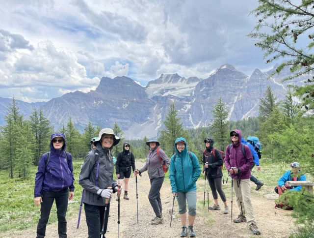A group of hikers in outdoor clothing and carrying trekking poles stand on a trail with mountains and trees in the background under a cloudy sky.