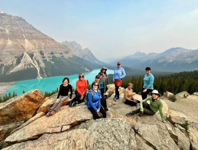 A group of people pose on rocky terrain overlooking a turquoise lake and forested mountains under a hazy sky.
