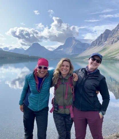 Three women in outdoor clothing stand smiling by a clear lake with mountains and clouds reflected in the water behind them.