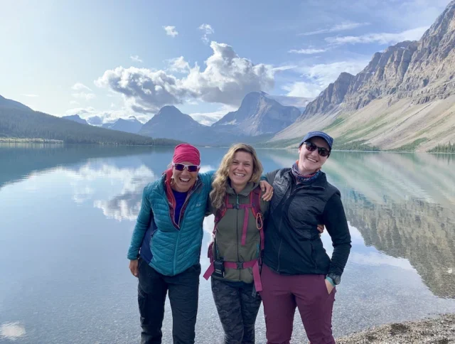 Three women in outdoor clothing stand smiling by a clear lake with mountains and clouds reflected in the water behind them.