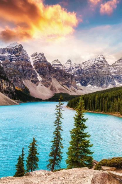 A turquoise lake surrounded by pine trees and rocky mountains under a partly cloudy, colorful sky at sunset.