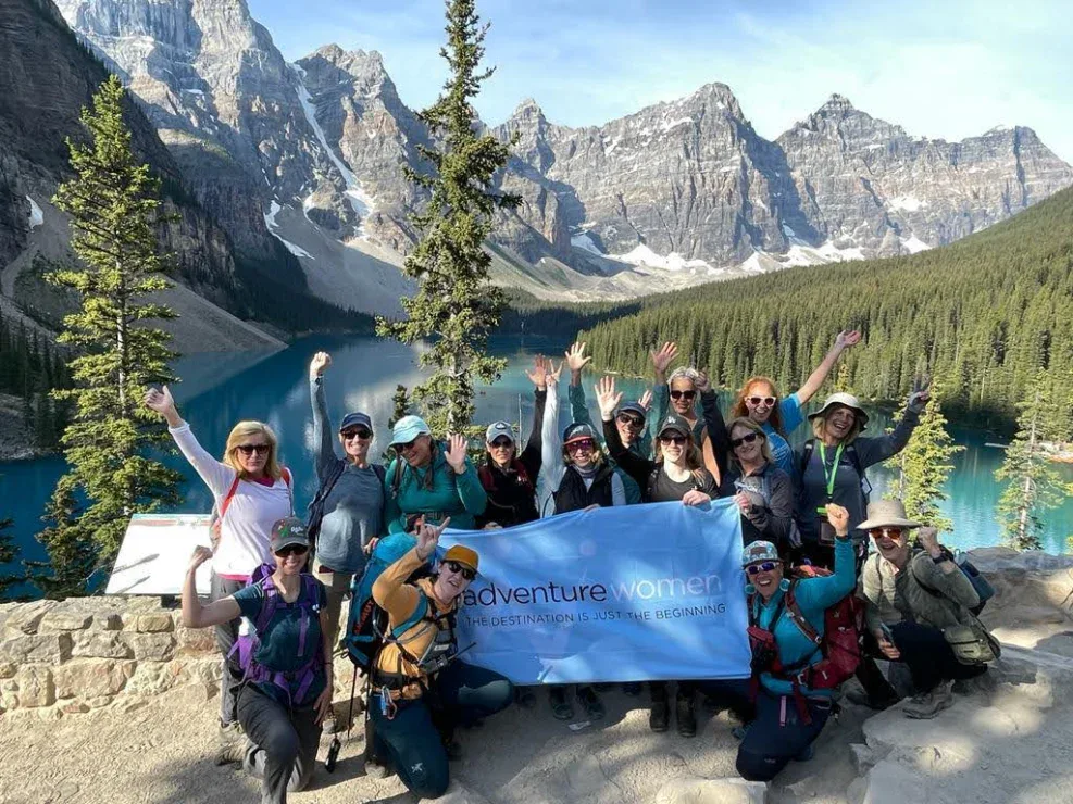 A group of women pose with a "Adventure Women" banner in front of a mountain lake and pine forest, with clear blue water and rocky peaks in the background.