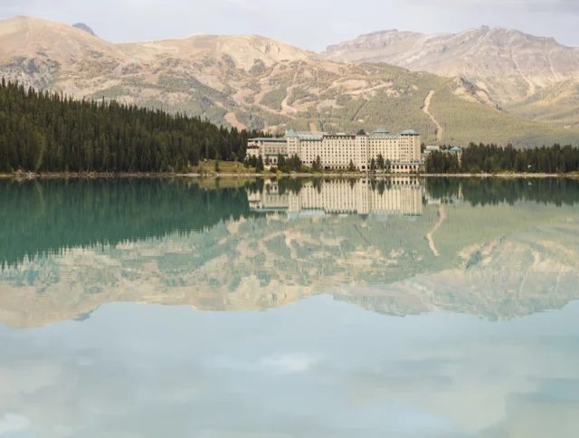 Large hotel building situated by a turquoise lake, surrounded by pine forest and mountains, with clear reflections in the calm water.