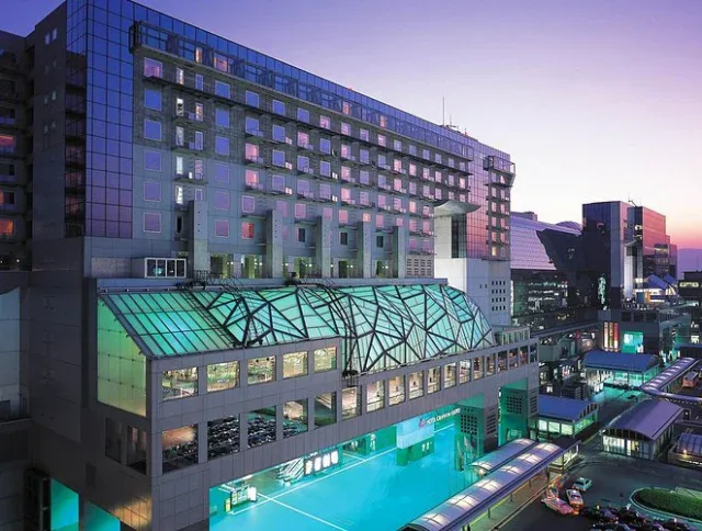 Modern multi-story building with large glass atrium, brightly lit at dusk, overlooking a busy urban area with adjacent structures and parked vehicles.