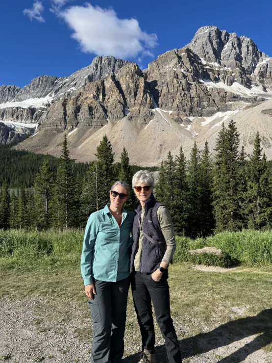 Two people stand together outdoors in front of pine trees and rocky, snow-dusted mountains under a blue sky with scattered clouds.