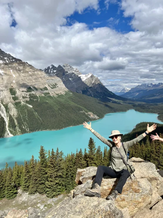 Person sitting on a rocky ledge with arms raised, overlooking turquoise lake, dense forest, and snow-capped mountains under a partly cloudy sky.