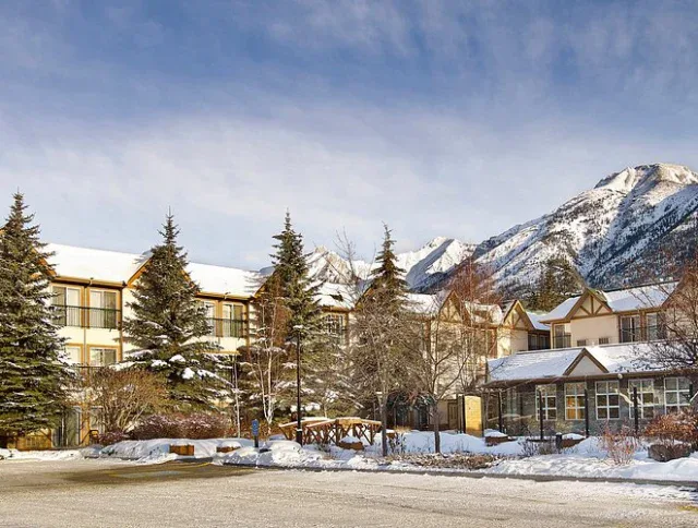 A lodge-style hotel with snow-covered roofs and pine trees stands at the base of a mountain range under a partly cloudy sky.