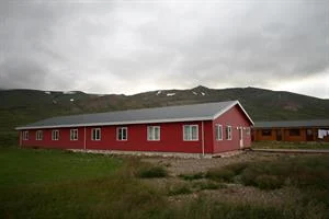 A long, single-story red building with white-framed windows sits on grass with mountains in the background under a cloudy sky.