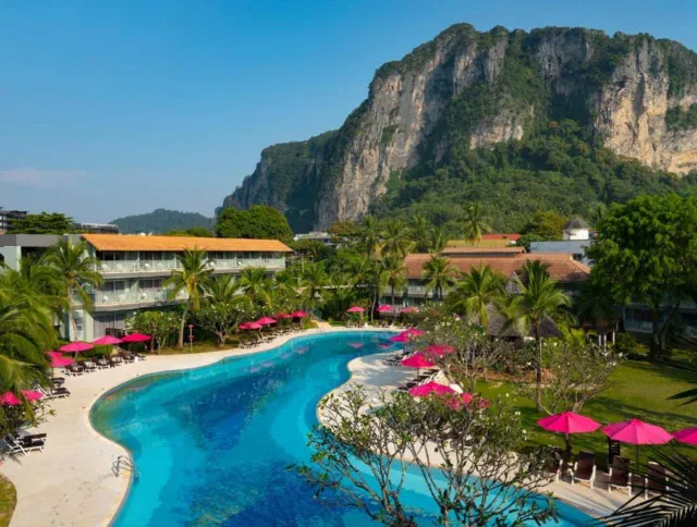Curved outdoor pool with pink umbrellas and palm trees, surrounded by buildings, set against a backdrop of large rocky cliffs and blue sky.