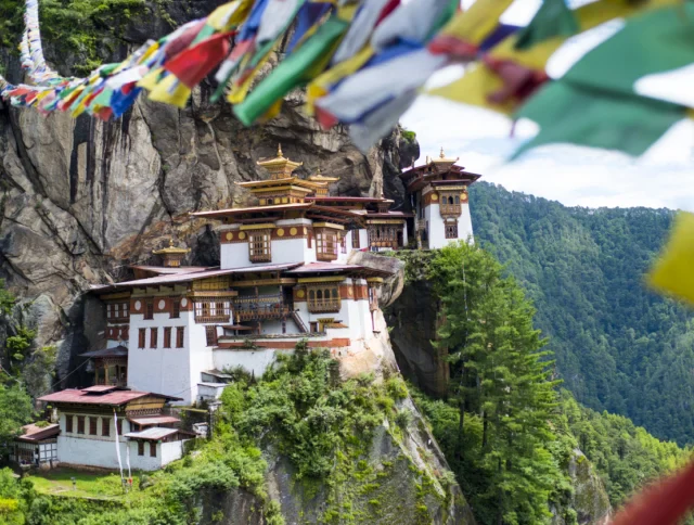 A monastery built on a cliffside surrounded by lush green forest, with colorful prayer flags in the foreground.