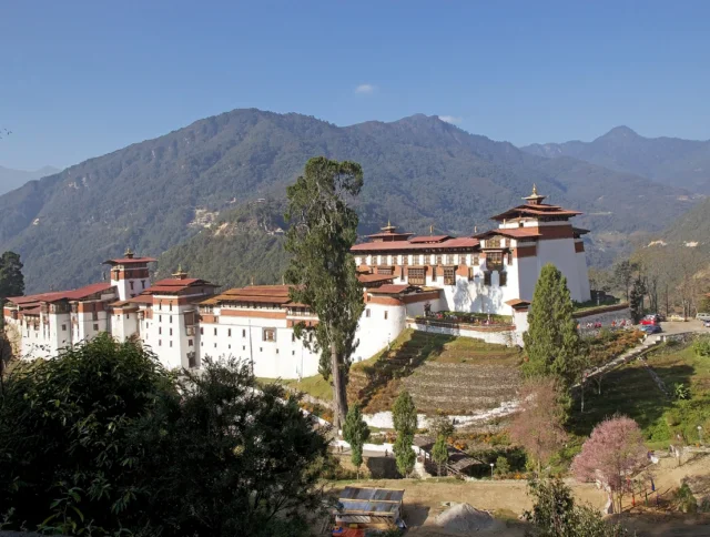 A large traditional fortress-monastery complex sits on a hillside, surrounded by green mountains and clear blue sky.