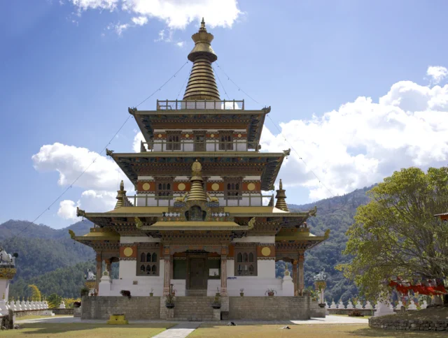 A traditional multi-tiered Buddhist temple with ornate details stands against a backdrop of hills and a partly cloudy sky.
