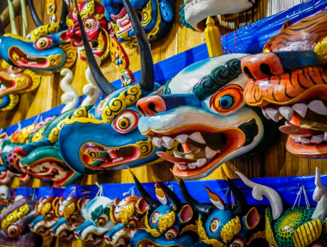 Colorful, intricately designed traditional dragon and animal masks are displayed in rows on a wooden wall.