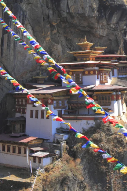 Tiger’s Nest Monastery in Bhutan sits on a cliffside, with colorful prayer flags strung in the foreground and forested mountains in the background.