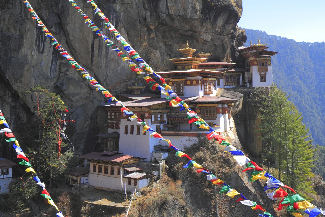Tiger’s Nest Monastery in Bhutan sits on a cliffside, with colorful prayer flags strung in the foreground and forested mountains in the background.