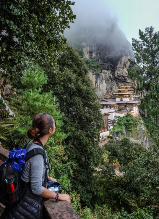 A person with a backpack stands on a path overlooking the Tiger's Nest Monastery built into a steep, forested cliffside, partially shrouded in mist.