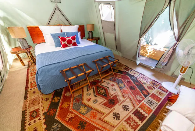 A bed with blue bedding and decorative pillows sits on a patterned rug in a tent with wooden side tables, lamps, a fan, and open flaps letting in natural light.