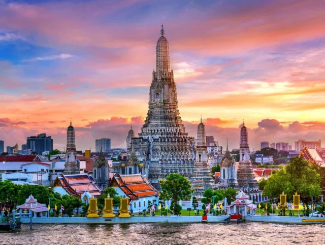 Wat Arun temple in Bangkok, Thailand, stands by the Chao Phraya River at sunset, with colorful sky and city buildings in the background.