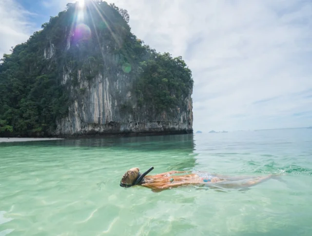 Person snorkeling in clear, shallow water near a small, lush island with steep cliffs under a partly cloudy sky and sunlight.