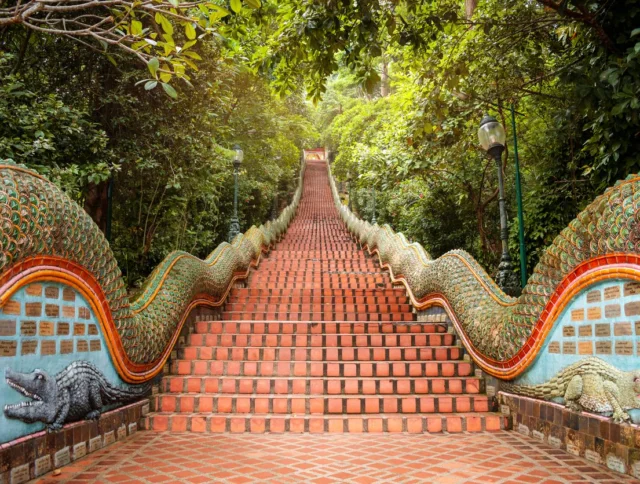 A long, red-tiled staircase flanked by ornate serpent sculptures leads upward through dense green foliage.