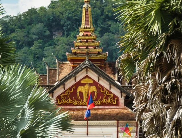 A traditional temple with ornate golden details and a pointed roof stands between palm trees, with a hill and cloudy sky in the background.