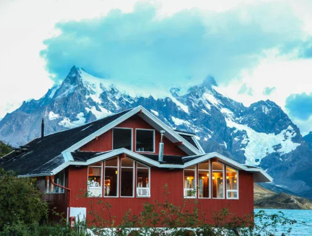 A red house with large windows sits in front of snow-capped mountains under a cloudy sky.